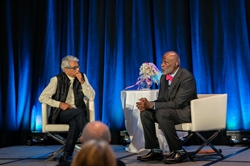 [ai] Two men seated on stage during an event, engaged in conversation. One man has gray hair and glasses, wearing a vest, while the other has a shaved head and is dressed in a suit with a bow tie. A table with flowers is in the background.